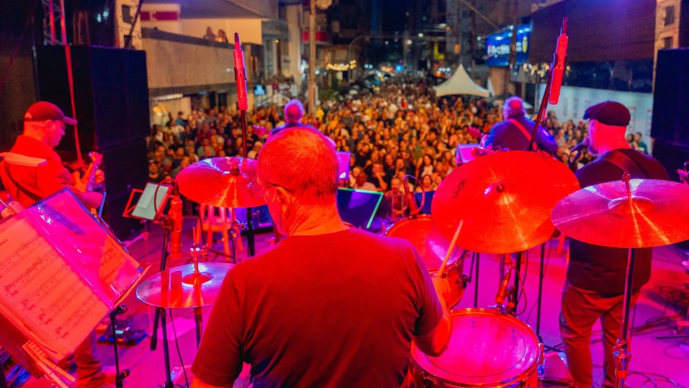 Músicos tocando instrumentos em palco iluminado em frente a uma multidão no Festival de Inverno de Guarapari 2025.